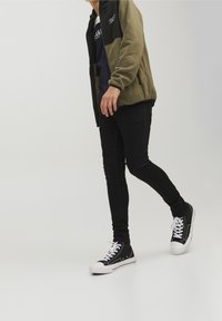 Veste en polaire vert olive avec un panneau supérieur noir, chemise bleu marine en dessous, jeans skinny noirs et baskets noires avec semelles blanches.