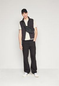 Svart pufferväst över en ljusbeige t-shirt, svarta byxor och vita sneakers med grå detaljer. Innehåller också en svart axelväska.