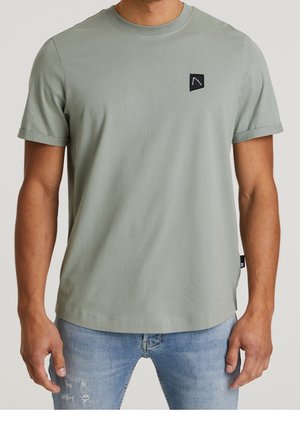 Hombre con camiseta de manga corta gris claro con un pequeño logo cuadrado negro en el pecho, combinado con vaqueros azul claro desgastados.