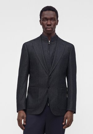 Homme portant un blazer gris foncé sur une veste intérieure charbon zippée, debout devant un fond clair uni.
