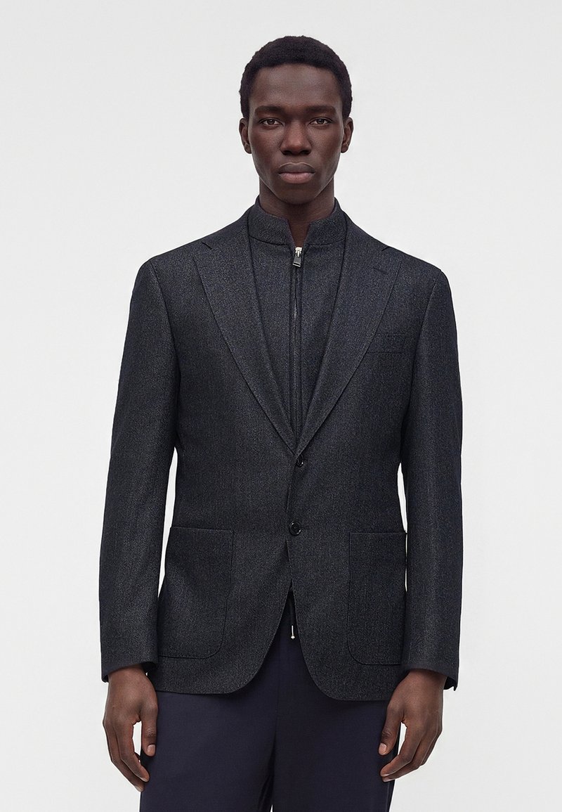 Homme portant un blazer gris foncé sur une veste intérieure charbon zippée, debout devant un fond clair uni.
