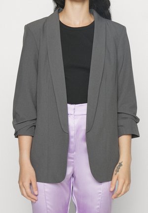 Personne portant un blazer gris foncé avec manches retroussées, chemise noire et pantalon en satin violet clair, mains détendues sur les côtés avec un tatouage de serpent visible au poignet droit.