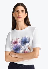 Haut blanc à manches courtes présentant un design floral dans des nuances de bleu, rose et violet, avec de subtils accents pailletés sur les fleurs.