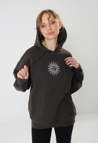 Smeđi oversized dukserica s grafikom sunca sprijeda. Ima kapuljaču s vezicom i rebrasti rub. Glatka tekstura i opušteni kroj.