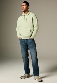 Light green pullover hoodie with a front pocket, paired with blue jeans and gray sneakers with white accents. Simple, casual design.