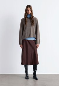 Brauner langer Rock, grauer flauschiger Pullover, blauer gerippter Rollkragenpullover. Schwarze kniehohe Stiefel, lockige Haare, neutraler Hintergrund.