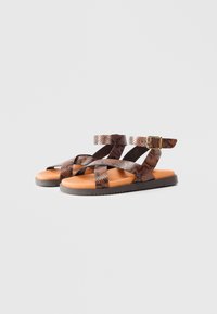 Sandalias de patrón de serpiente marrón con tiras cruzadas, hebilla ajustable en el tobillo y plantilla acolchada color cuero sobre una suela de goma negra.