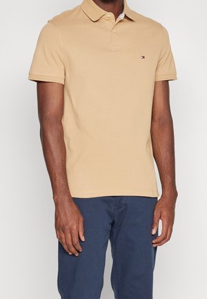 Man draagt een beige polo met korte mouwen en een klein logo op de borst, gecombineerd met marineblauwe broek, staand tegen een effen witte achtergrond.