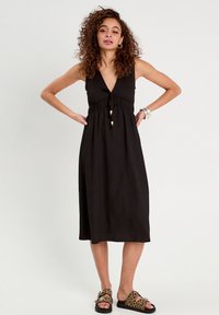 Robe noire sans manches avec un décolleté en V, une taille smockée et des accents en perles en bois. Le mannequin porte des sandales à imprimé léopard. Tissu lisse.