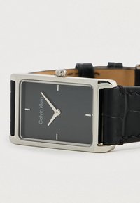 Montre en quartz carrée avec un cadran noir, des aiguilles argentées et un bracelet en cuir noir avec motif crocodile. Logo affiché à 12 heures.