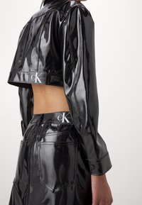 Black glossy faux leather cropped jacket with long sleeves, featuring a logo patch and matching high-waisted skirt with back pockets.