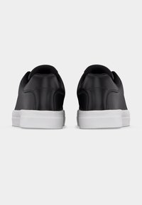 Ζευγάρι μαύρα χαμηλά αθλητικά παπούτσια με λευκές λαστιχένιες σόλες, εμφανιζόμενα από πίσω σε απλό λευκό φόντο.