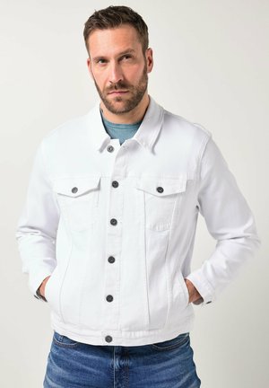 Bärtiger Mann mit kurzem braunem Haar, der eine weiße, geknöpfte Jeansjacke über einem türkisfarbenen Hemd trägt, Hände in den Taschen, steht vor einem einfarbigen Hintergrund.