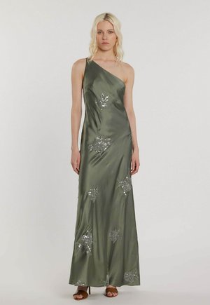 Donna bionda che indossa un abito di raso verde oliva lungo fino a terra con una spalla scoperta e dettagli floreali in paillettes argento, in piedi su uno sfondo semplice.