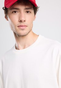Weißes Crewneck-Sweatshirt mit weicher Textur, präsentiert mit einer roten Kappe. Verfügt über eine einfache, lockere Passform und ein minimalistisches Design.