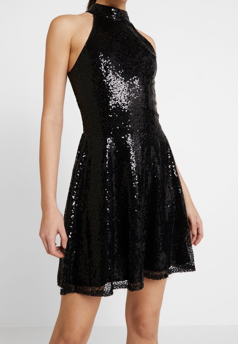 Ärmlös svart paljettklänning med halterneck, med figurnära överdel och utsvängd kjol, bärs av en stående person mot en enhetlig bakgrund.