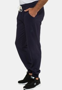 Marineblaue Sweatpants mit elastischem Bund und Kordelzug, ausgestattet mit Seitentaschen und einem kleinen Logo, kombiniert mit schwarzen Sportschuhen.