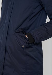 Persona con chaqueta azul oscuro, con la mano parcialmente dentro del bolsillo lateral con cierre de botón a presión y puño negro visible.