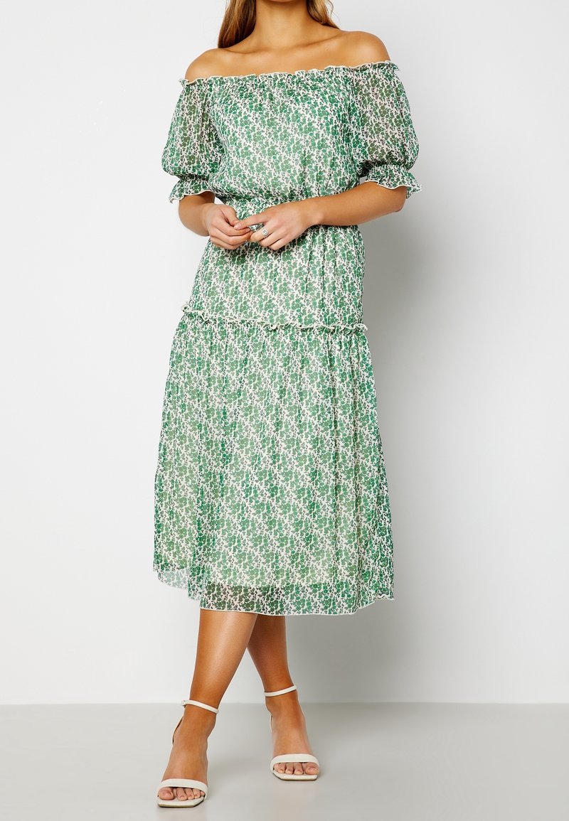 Femme portant une robe midi verte et blanche à fleurs, épaules dénudées, manches bouffantes et bords volantés, associée à des talons blancs à bride cheville.