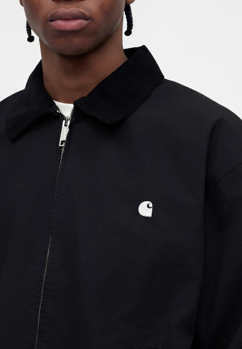 Nahaufnahme einer Person, die eine schwarze Jacke mit Reißverschluss und einem Kordkragen sowie einem kleinen weißen Logo auf der linken Brust trägt.