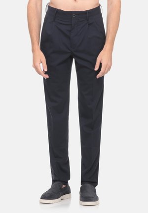 Uomo con pantaloni aderenti neri e scarpe scure slip-on, in piedi davanti a uno sfondo bianco semplice.