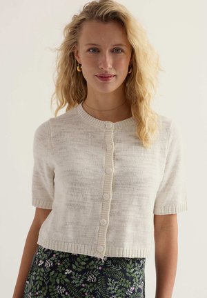 Šviesiaplaukė moteris, dėvinti trumpomis rankovėmis kremišką megztą kardiganą su sagomis ir žalią gėlėtą sijoną ant vienspalvio fono.