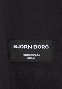 Črn material z pravokotno oznako, na kateri so v belem napisi "BJÖRN BORG," "STOCKHOLM," in "1956," sešiti v kotiček.