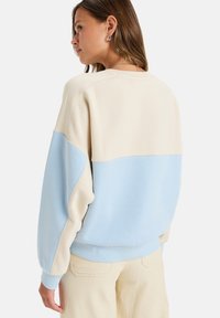 Sweatshirt color-blocké beige clair et bleu ciel avec une coupe décontractée, des coutures visibles et des poignets côtelés. Texture de tissu douce.