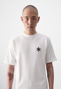 T-shirt en coton blanc avec des manches courtes et un col rond. Logo astérisque noir sur la poitrine gauche, texture lisse.