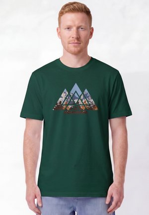 Mann trägt ein dunkelgrünes T-Shirt mit einer Grafik von überlappenden Bergformen, die mit Wald- und Himmelmustern gefüllt sind.
