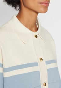 Sweter dziany z kołnierzem, w kolorze kremowym z jasnoniebieskimi poziomymi paskami i złotymi guzikami przy kołnierzu.