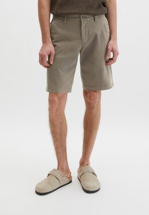 Mann trägt hellbraune kniehohe Shorts und beige Slipper, steht vor einem schlichten weißen Hintergrund.