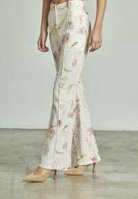 Vrouw in hoge taille, met bloemenpatroon en brede pijpen, met beige puntige hakken, staande op een betonnen vloer.