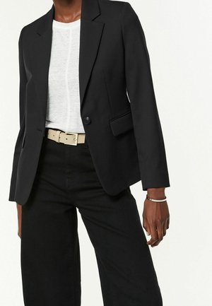 Personne portant un blazer noir, un t-shirt blanc, un pantalon noir, une ceinture beige et un bracelet fin au poignet droit.