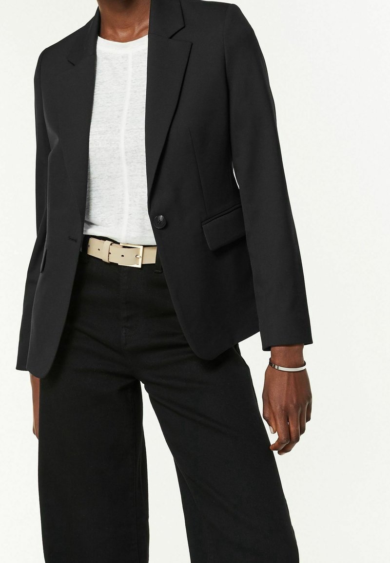 Personne portant un blazer noir, un t-shirt blanc, un pantalon noir, une ceinture beige et un bracelet fin au poignet droit.