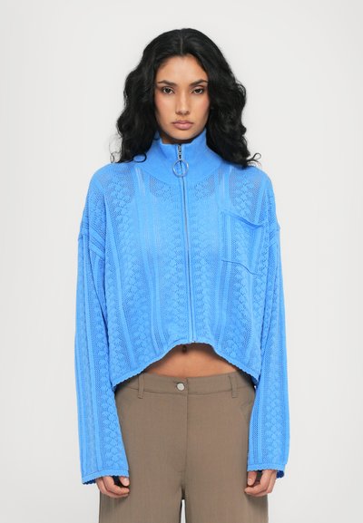 Sweat à zip cropped bleu clair avec un col montant, doté d'une tirette de zip circulaire et d'un motif de tricot texturé. Manches longues, coupe décontractée.