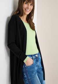 Femme souriante, portant un haut vert clair, un cardigan noir et un jean bleu, avec une main dans la poche, appuyée contre un mur blanc.