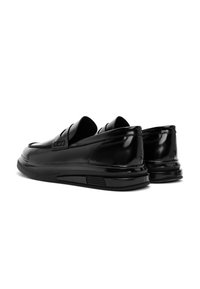 Une paire de mocassins en cuir noir brillant avec des semelles en caoutchouc, inclinée pour montrer les côtés extérieur et arrière sur un fond blanc.