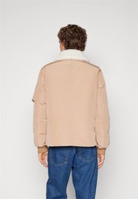 Beige puffjacka med en vit fårskinnskrage, elastiska ärmslut och en avslappnad passform, visad bakifrån mot en neutral bakgrund.