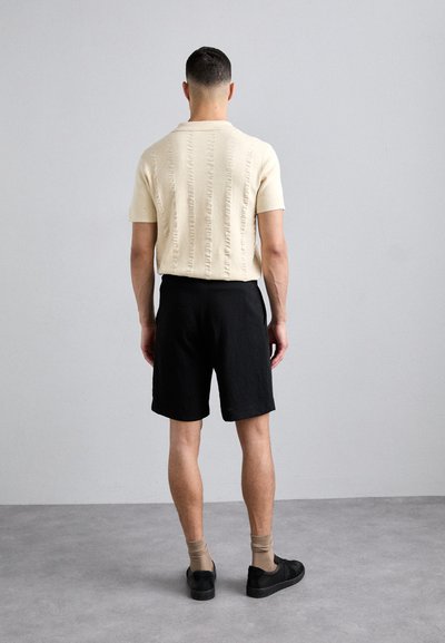 Lysbeige striktrøje med kortærmer og strukturerede striber, parret med sorte shorts. Model iført brune sokker og sorte sneakers.