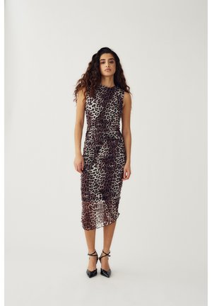 Femme aux cheveux longs et bouclés portant une robe midi à imprimé léopard sans manches et des talons pointus noirs, posant devant un fond uni.