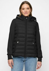 Woman wearing a black quilted jacket with a hood and zipped pockets, paired with light blue jeans, standing against a plain white background.