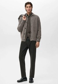 Cardigan de lana gris sobre una camisa oscura con botones, combinado con pantalones oscuros y zapatos negros. El modelo posa con una mano en el hombro del cardigan.