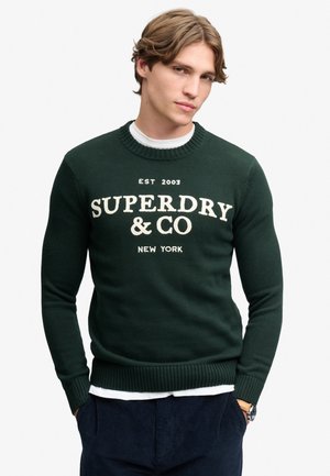 Young man with light brown hair wearing a dark green Superdry sweatshirt and dark pants, standing with hands in pockets.