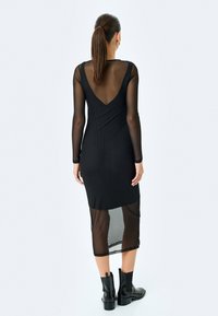 Długa czarna sukienka z przezroczystymi rękawami i tyłem, dopasowana sylwetka, dekolt w serek i długość do połowy łydki, zestawiona z czarnymi botkami za kostkę.