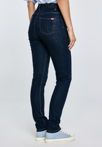 Inimene, kes kannab tumelau siniseid kitsaid teksapükse ja helesiniseid tossusid, seisab käega külje peal.