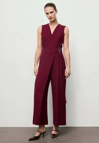 Burgundský overál s překládaným designem, výstřihem do V, bez rukávů a stříbrnou sponou v pase. Široké nohavice a strukturovaný materiál.