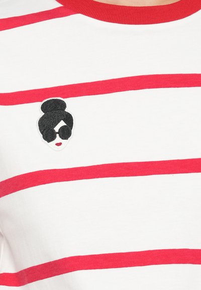 T-shirt branca com riscas horizontais vermelhas, apresentando um rosto bordado em preto e branco a usar óculos de sol no lado esquerdo.