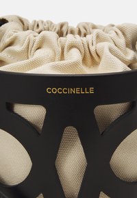 Crna kožna torba s bež tkaninom na vrhu, s izrezanom dizajnom i zlatnim "Coccinelle" brendiranjem. Teksture materijala vizualno se kontrastiraju.
