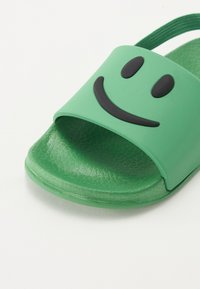 Grønn glidesandal laget av glatt plast, med et markant svart smilefjesgrafikk på stroppen og en teksturert såle.